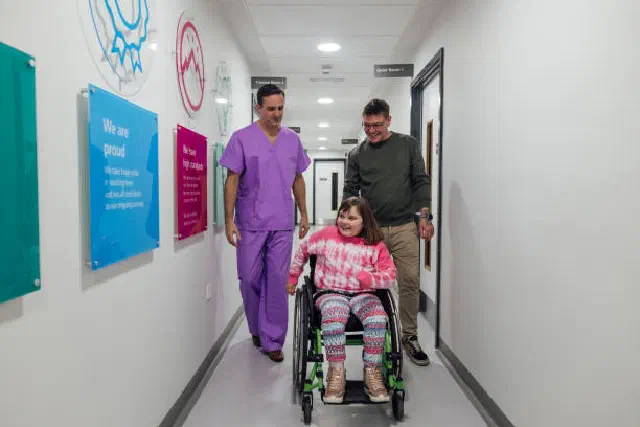 A patient using a wheelchair accompanied by a caregiver and healthcare professional